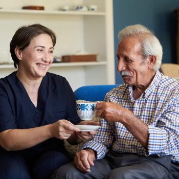Professional helpful caregiver at nursing home. Health visitor and a senior man during home visit. Young caregiver in uniform and elderly man drinking tea